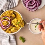 A bowl of Kay's Curries Tofu Biryani with tofu and green herbs, topped with red onion rings, shown on a beige surface with a hand holding a spoonful of the rice. A small bowl of white sauce and a plate of sliced red onion are also visible.
